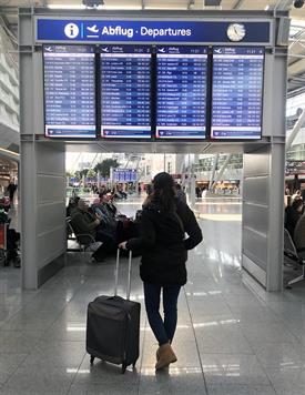 Am Flughafen. Es ist eine große blaue Tafel mit Abflugzeiten zu sehen. Davor steht eine junge Frau mit dunklen Haaren, dunkler Jacke und einem kleinen Koffer und blickt auf die Tafel.